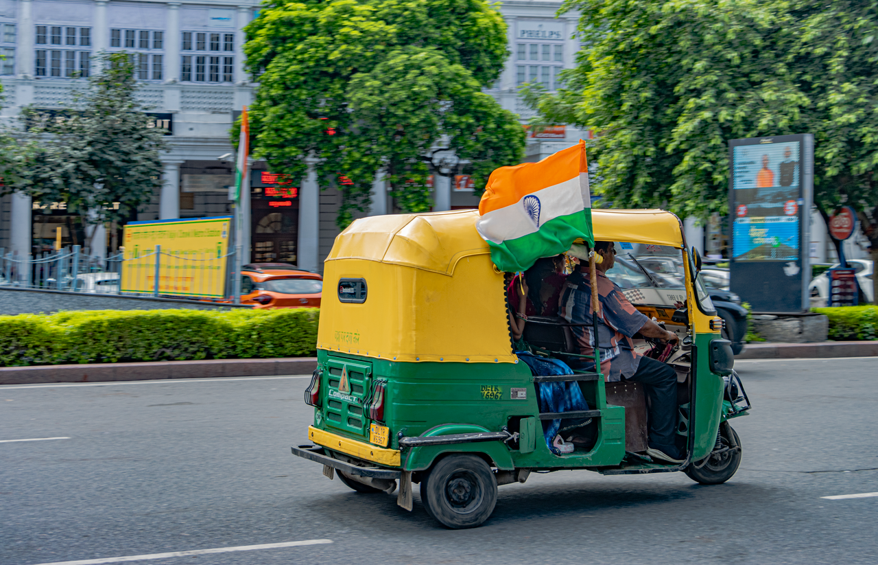 A green and yellow auto rickshaw adorned with the Indian flag drives through New Delhi streets.